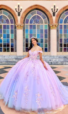A young woman smiles in front of arched windows and columns, wearing the Quinceanera Dress PR30162 by Princesa—a lavender ball gown with floral details and an off-the-shoulder neckline.
