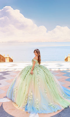 A woman in the Quinceanera Dress PR30162 By Princesa, featuring an ornate pastel ombre design and off-the-shoulder neckline, stands on a patterned terrace overlooking a calm body of water beneath a partly cloudy sky.
