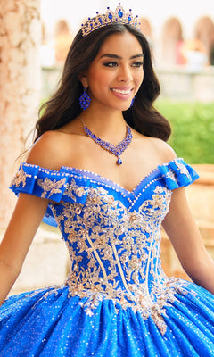 A young woman stands outdoors, smiling in the Quinceanera Dress PR30163 By Princesa—a jeweled tiara atop her head and an ornate blue and gold lace ball gown with a cathedral train.