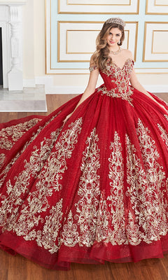 A woman wears the Quinceanera Dress PR30171 By Princesa, a red ball gown with gold embroidery and layers of glitter tulle, paired with a tiara. She sits indoors on a wooden floor before a decorated wall and fireplace.