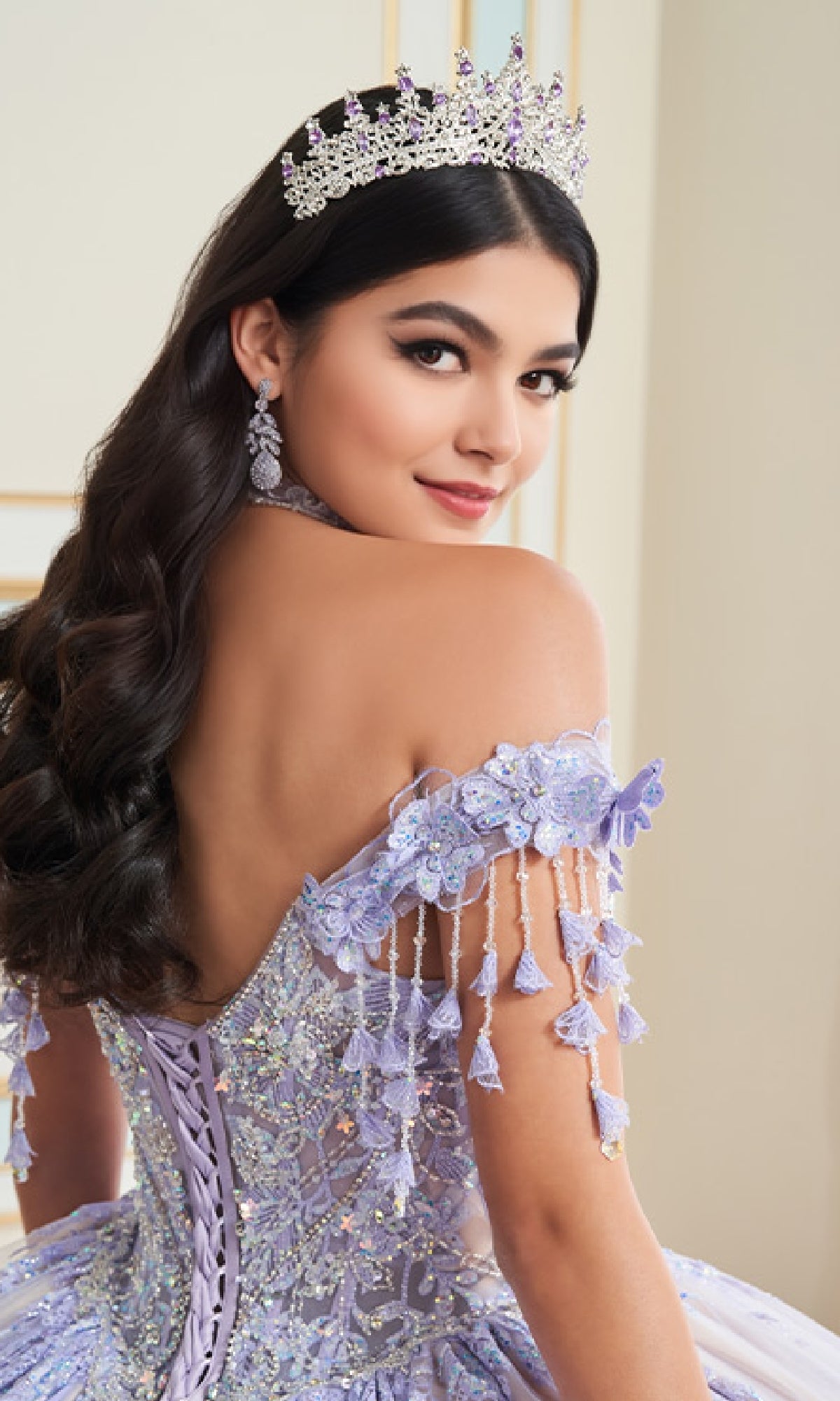 A young woman with long dark hair wears the Princesa PR30172 Glow-in-the-Dark Quinceanera Dress, an off-shoulder lavender gown with lace, beading, and 3D floral appliques, looking over her shoulder at the camera.