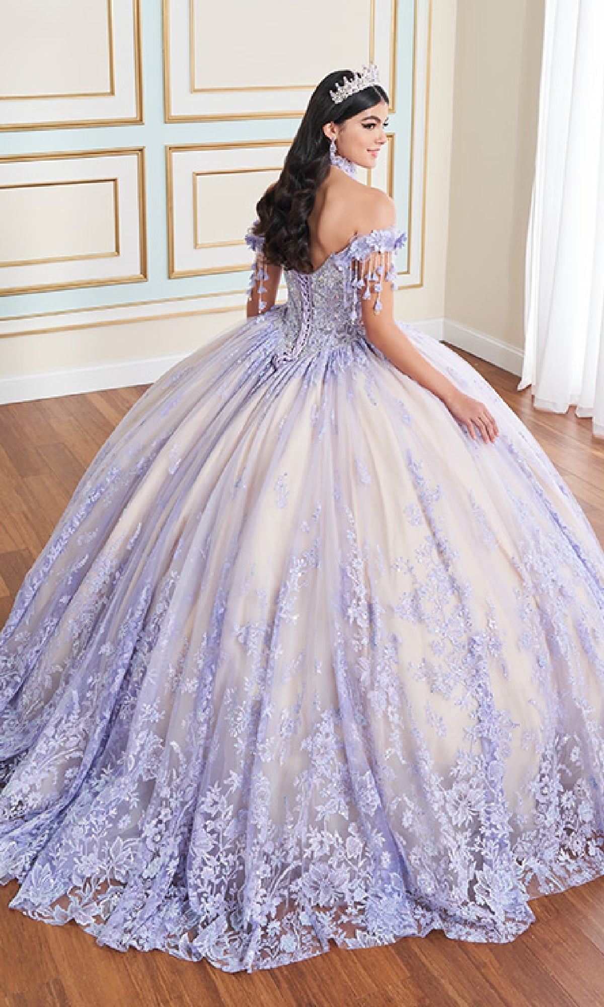 A woman stands indoors, facing away, wearing the Princesa PR30172 glow-in-the-dark Quinceanera dress in lavender and white with 3D floral appliques and lace, a tiara atop her long wavy dark hair.