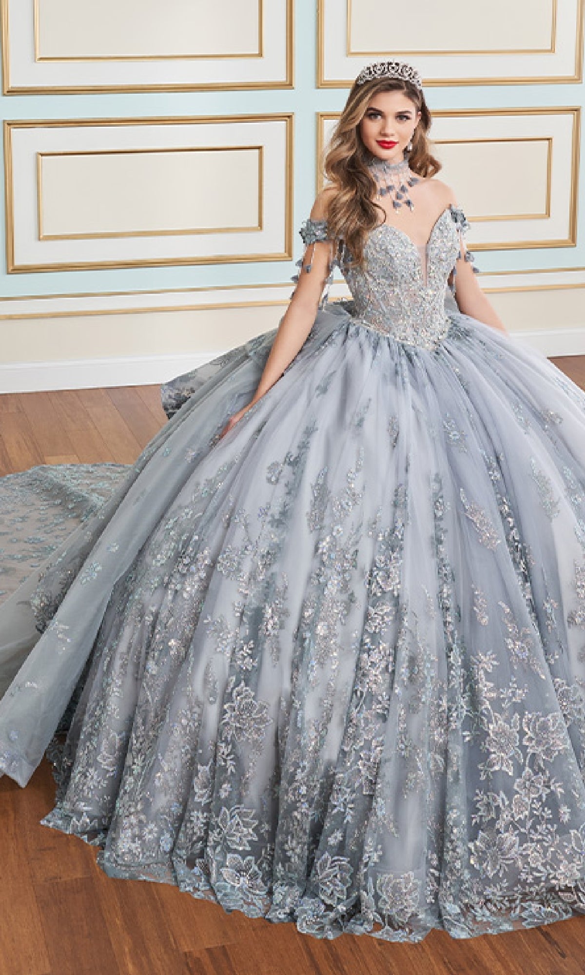 A woman in the Glow-in-the-Dark Quinceanera Dress: Princesa PR30172, featuring a light blue hue, delicate beading, 3D floral appliques, a long train, and a tiara, stands indoors against an ornate wall.
