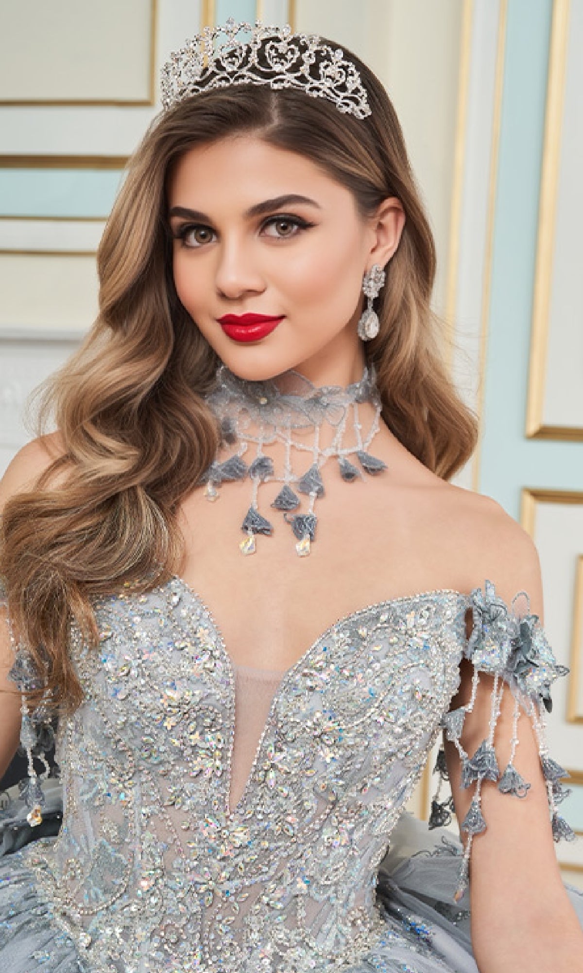 A woman poses indoors wearing the Glow-in-the-Dark Quinceanera Dress: Princesa PR30172, featuring a jeweled tiara and a sparkling off-shoulder silver gown with intricate beading and 3D floral appliques.