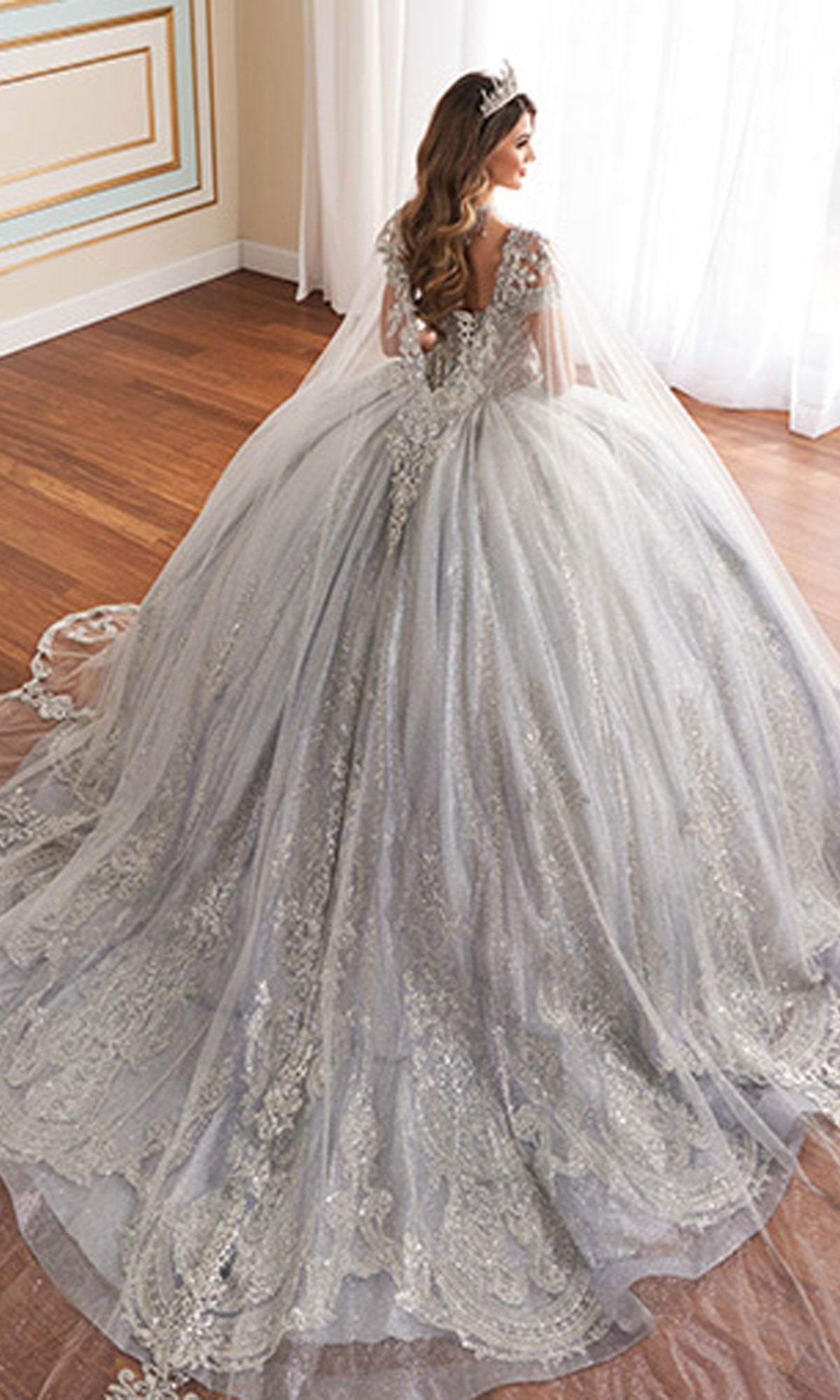 A woman wearing the Quinceanera Dress PR30173 By Princesa—a silver, intricately detailed off-the-shoulder gown with a long train—stands by a sunlit window, crowned and veiled.