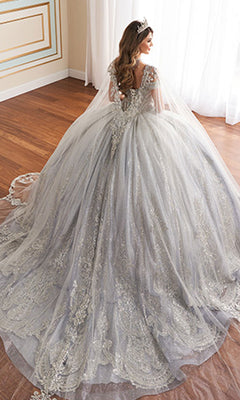 A woman wearing the Quinceanera Dress PR30173 By Princesa—a silver, intricately detailed off-the-shoulder gown with a long train—stands by a sunlit window, crowned and veiled.