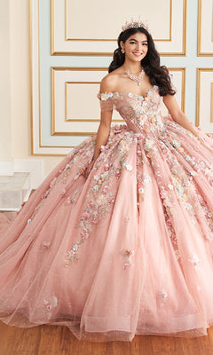 A woman in the Quinceanera Dress PR30175 By Princesa, a pink off-the-shoulder ball gown with beading and 3D floral details, wears a tiara and stands in an ornate room with gold-trimmed walls.