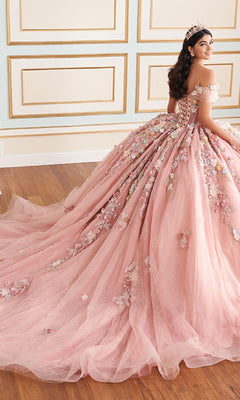 A young woman in the Quinceanera Dress PR30175 By Princesa, featuring off-shoulder 3D pink florals and delicate beading, wears a tiara as she sits in an elegant room with paneled walls and wooden floors.