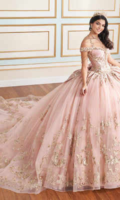 A woman wears the Quinceanera Dress PR30176 By Princesa, a pink ball gown with gold embroidery, metallic beading, a detachable back bow, and long train, standing on a wooden floor before a cream wall with gold trim.