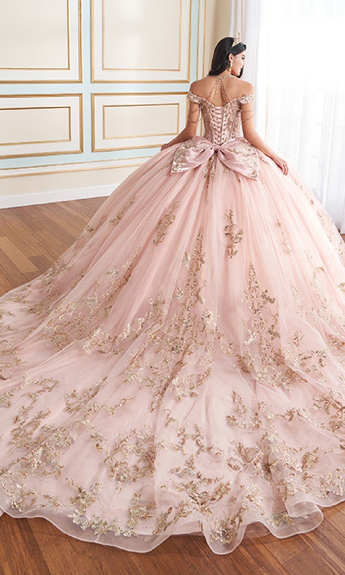 A woman in the Quinceanera Dress PR30176 By Princesa, an off-the-shoulder pink ball gown with gold embroidery, metallic beading, and a large detachable back bow, stands facing away in an elegant, bright room.
