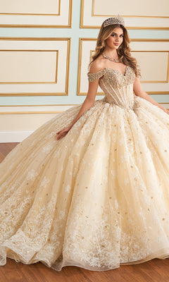 A woman in the Quinceanera Dress PR30177 By Princesa, featuring an ornate off-the-shoulder corset bodice and full skirt, with a jeweled tiara and necklace, stands in an elegant room with paneled walls.