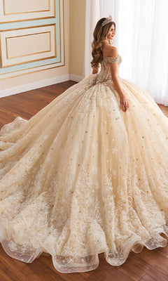 A woman smiles and stands on a wooden floor wearing the Quinceanera Dress PR30177 By Princesa, an ornate off-the-shoulder gown with a corset bodice and lace embroidery. Her tiara catches the light as she faces slightly away.