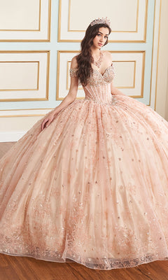 A woman sits indoors against a decorated wall wearing the Quinceanera Dress PR30177 By Princesa, featuring an off-the-shoulder corset bodice with pink embellishments, and finishes her look with a sparkling tiara.