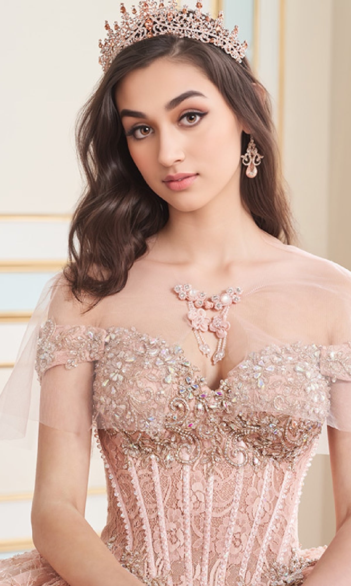 A young woman poses indoors wearing jeweled accessories and the Quinceanera Dress PR30177 By Princesa, an off-the-shoulder gown with a corset bodice, lace embroidery, and beadwork.