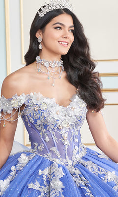 A woman wearing the Princesa Quinceanera Dress PR30178—a blue, off-the-shoulder gown with a corset bodice, floral appliqués, and beadwork—smiles indoors with a jeweled tiara.