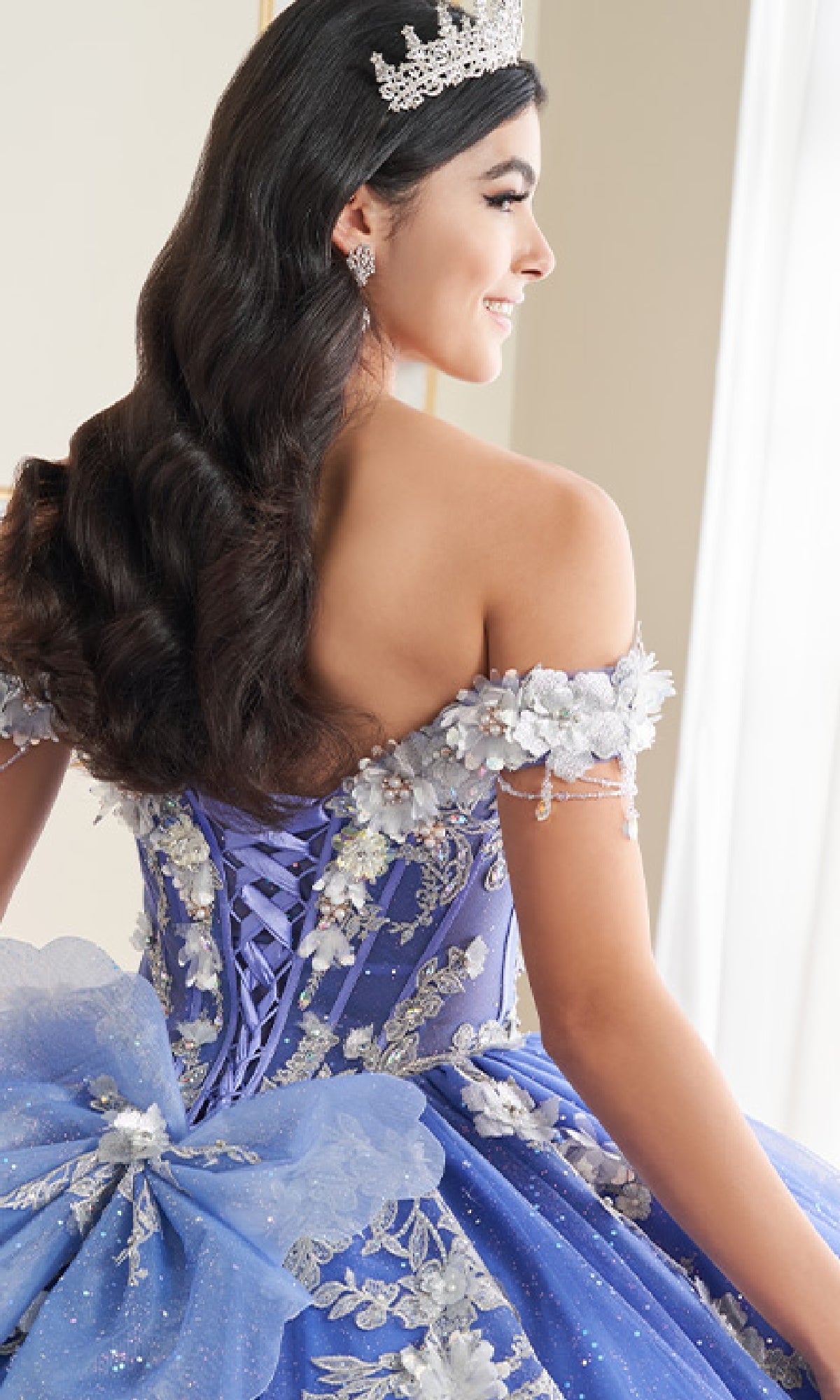 A young woman viewed from behind wears the Quinceanera Dress PR30178 By Princesa, featuring a tiara, detailed blue fabric, floral decorations, and a corset bodice.