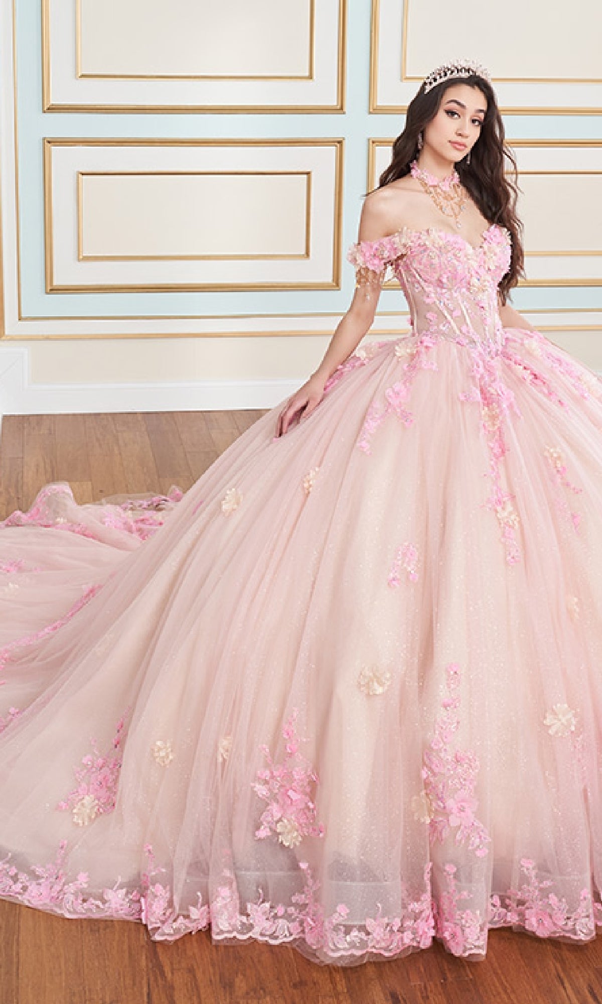A young woman in the Quinceanera Dress PR30178 By Princesa, featuring glitter tulle, floral details, a corset bodice, and a tiara, poses in an elegant room with wood floors and decorative wall panels.