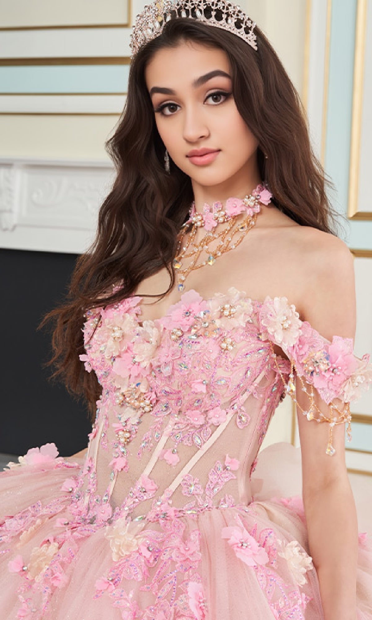A young woman wears the Quinceanera Dress PR30178 By Princesa, featuring a jeweled tiara, off-shoulder pink floral design, corset bodice, intricate embroidery, and beadwork as she stands indoors.
