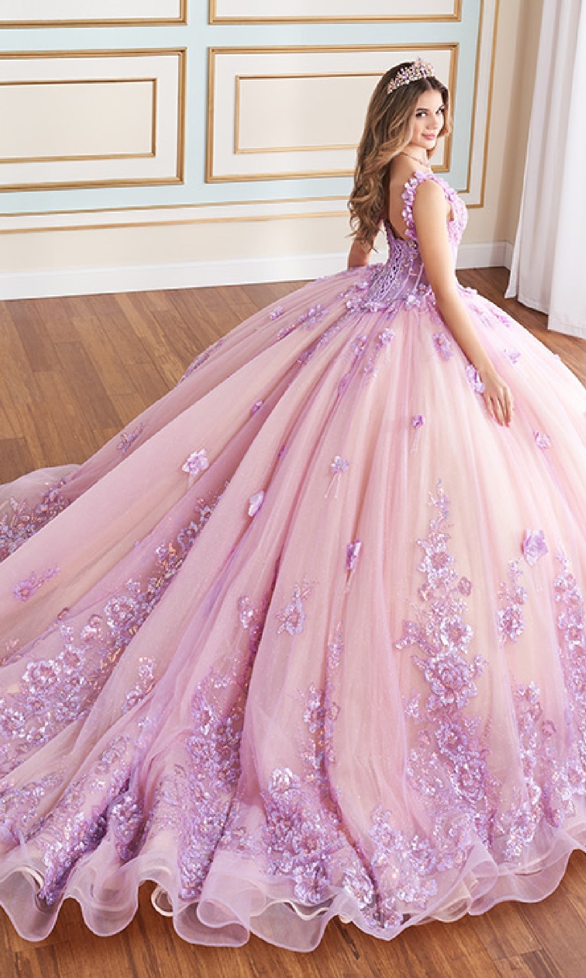A young woman wearing the Quinceanera Dress PR30179 by Princesa, adorned with purple 3D flowers and a tiara, stands on a wooden floor, looking over her shoulder.