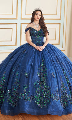 A woman in the Quinceanera Dress PR30179 by Princesa—a sparkling navy blue ball gown with off-the-shoulder sleeves, glitter tulle, 3D flowers, and a tiara—stands indoors against a paneled wall.