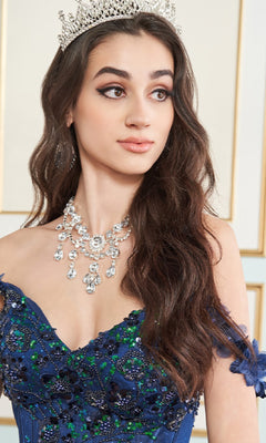 A young woman in the Quinceanera Dress PR30179 By Princesa, featuring an embellished blue ball gown with glitter tulle, jeweled crown, and necklace, looks to the side.