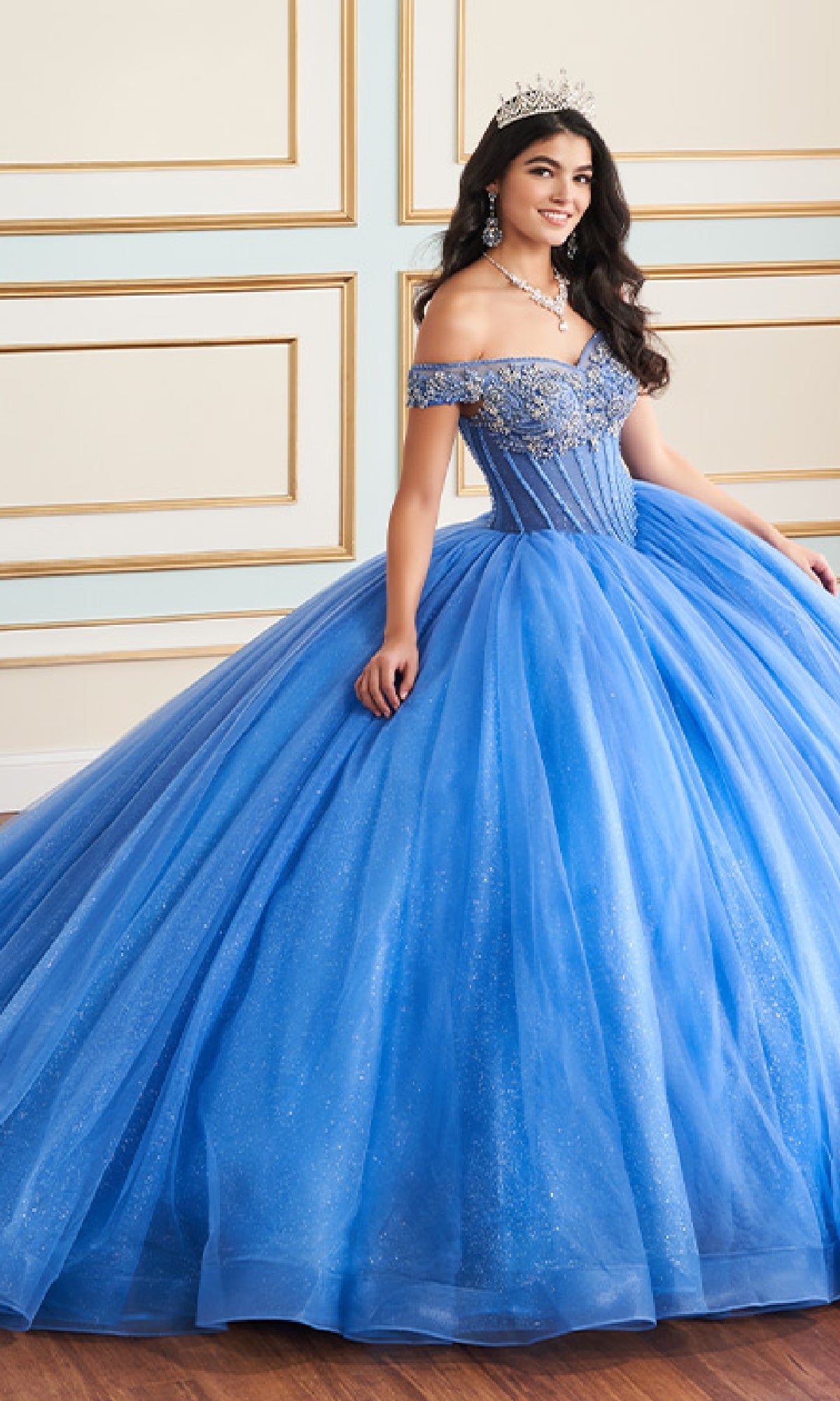 A woman in the Quinceanera Dress PR30180 by Princesa, featuring a blue off-the-shoulder beaded bodice and glitter tulle full skirt, stands in an elegant room adorned with a tiara and jewelry.