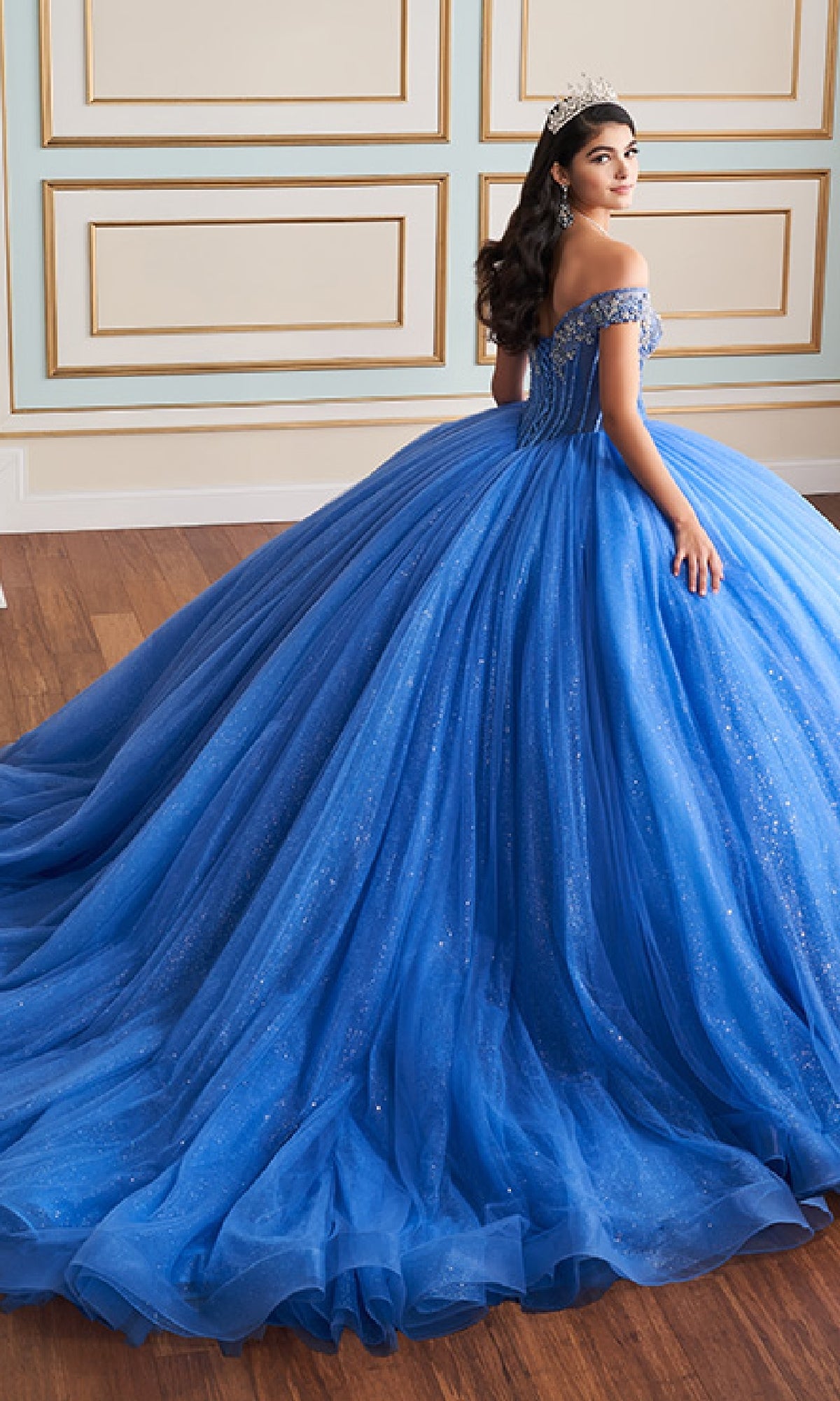 A young woman in the Quinceanera Dress PR30180 By Princesa, a glitter tulle ball gown with an off-the-shoulder neckline and a tiara, stands in a formal room with wood floors and decorative wall paneling.