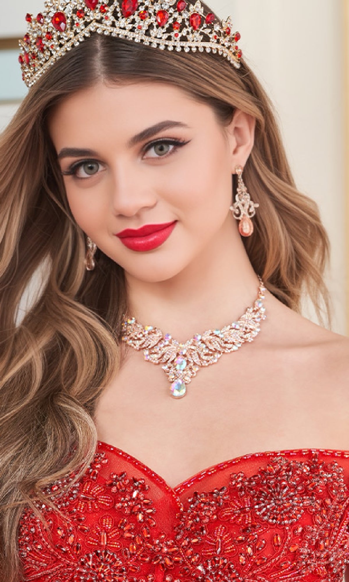 A young woman wearing the Quinceanera Dress PR30180 By Princesa—a glitter tulle ball gown with sparkling beaded embellishments—paired with a jeweled tiara, chandelier earrings, and a statement necklace, looking at the camera.