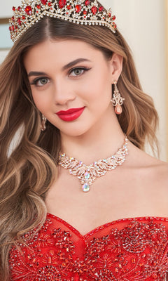 A young woman wearing the Quinceanera Dress PR30180 By Princesa—a glitter tulle ball gown with sparkling beaded embellishments—paired with a jeweled tiara, chandelier earrings, and a statement necklace, looking at the camera.