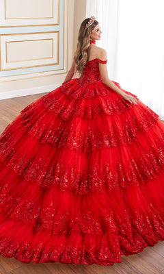 A woman in the Quinceanera Dress PR30181 By Princesa, featuring sparkling tiers and off-the-shoulder sleeves with a tiara, stands in a decorated room, facing away from the camera.