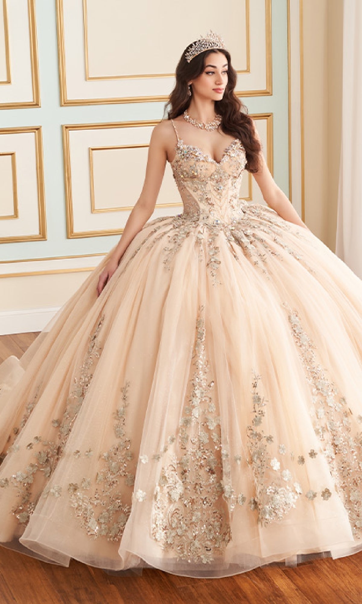A woman stands indoors near a decorative wall wearing the Quinceanera Dress PR30182 By Princesa—a tiara-topped, ornate light peach ball gown with floral embellishments and glitter tulle.