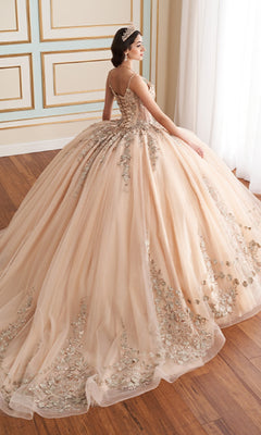 A woman wearing the Quinceanera Dress PR30182 by Princesa, a peach ball gown with silver floral details and glitter tulle layers, sits on a wooden floor near a window.