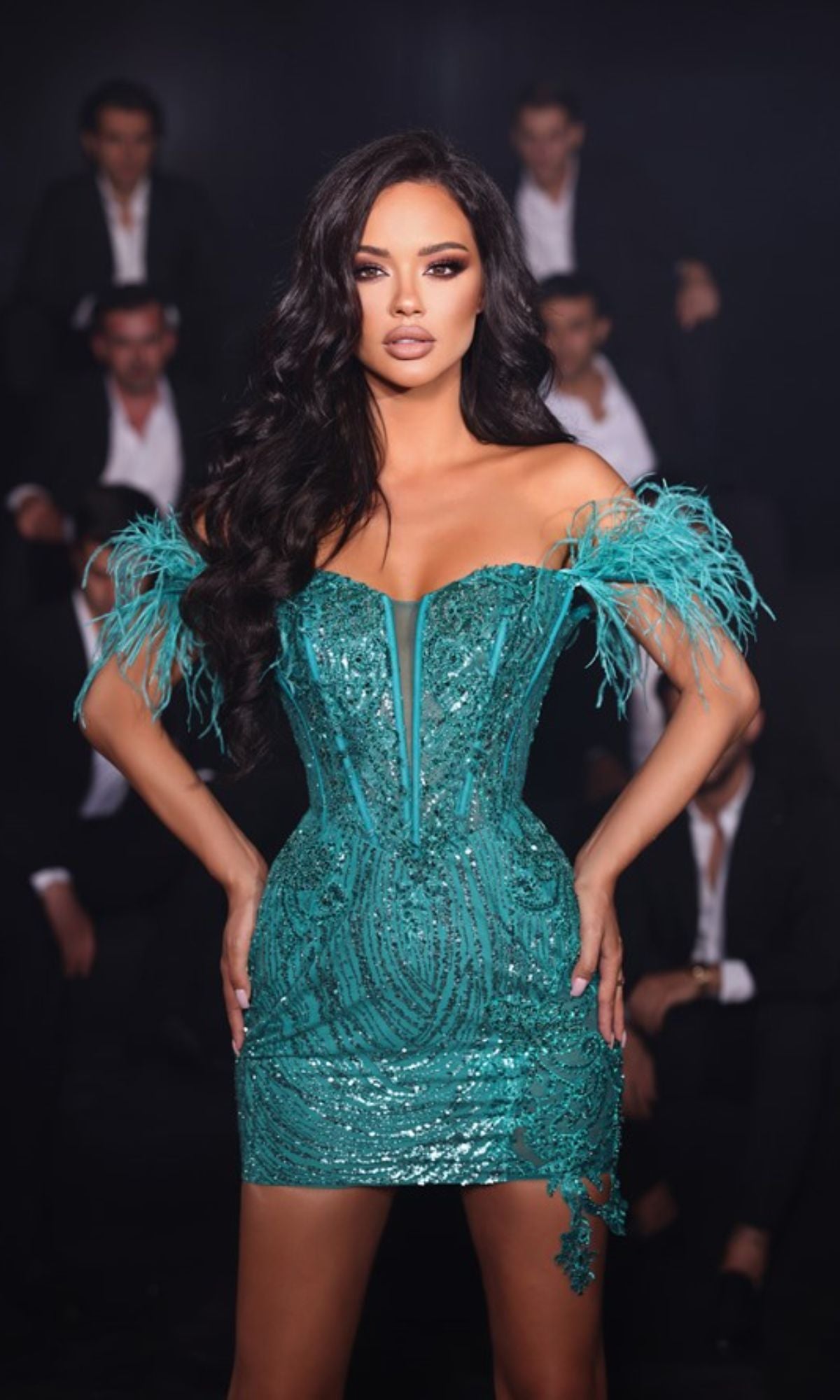 A woman confidently wears the Short Party Dress PS23147 by Portia and Scarlett, featuring teal sequins and off-the-shoulder feathered sleeves, while several men in suits sit in the dimly lit background.