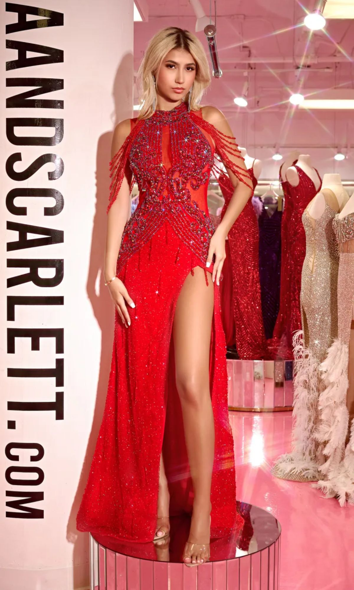 A woman wearing the red Long Prom Dress PS23704C by Portia and Scarlett stands in a store beside mannequins dressed in similar sheer illusion gowns.