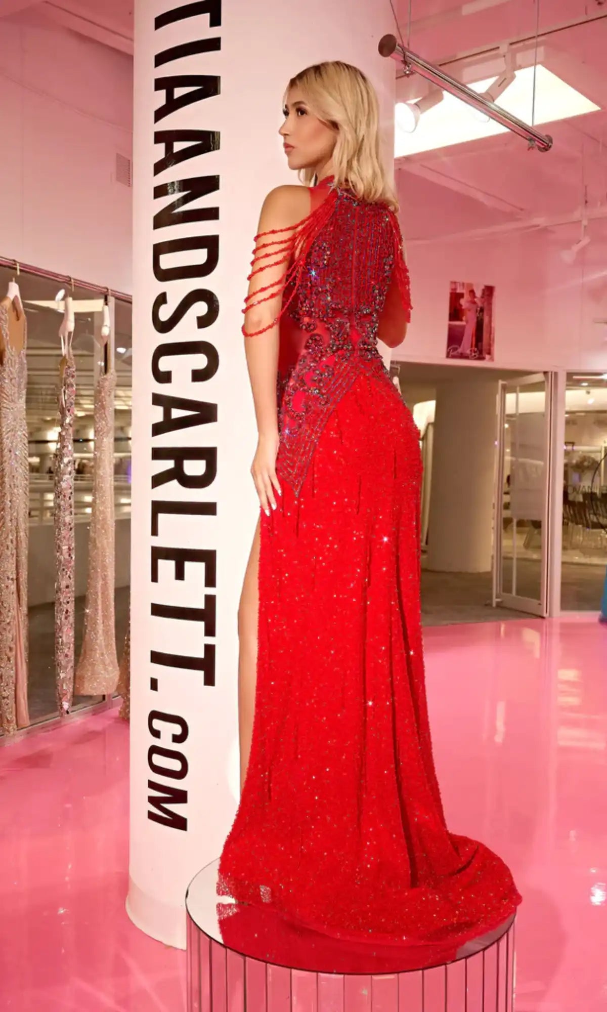 A woman stands on a platform in a brightly lit showroom, facing away from the camera, wearing the Long Prom Dress PS23704C by Portia and Scarlett—a sparkling red beaded formal gown with sheer illusion details.
