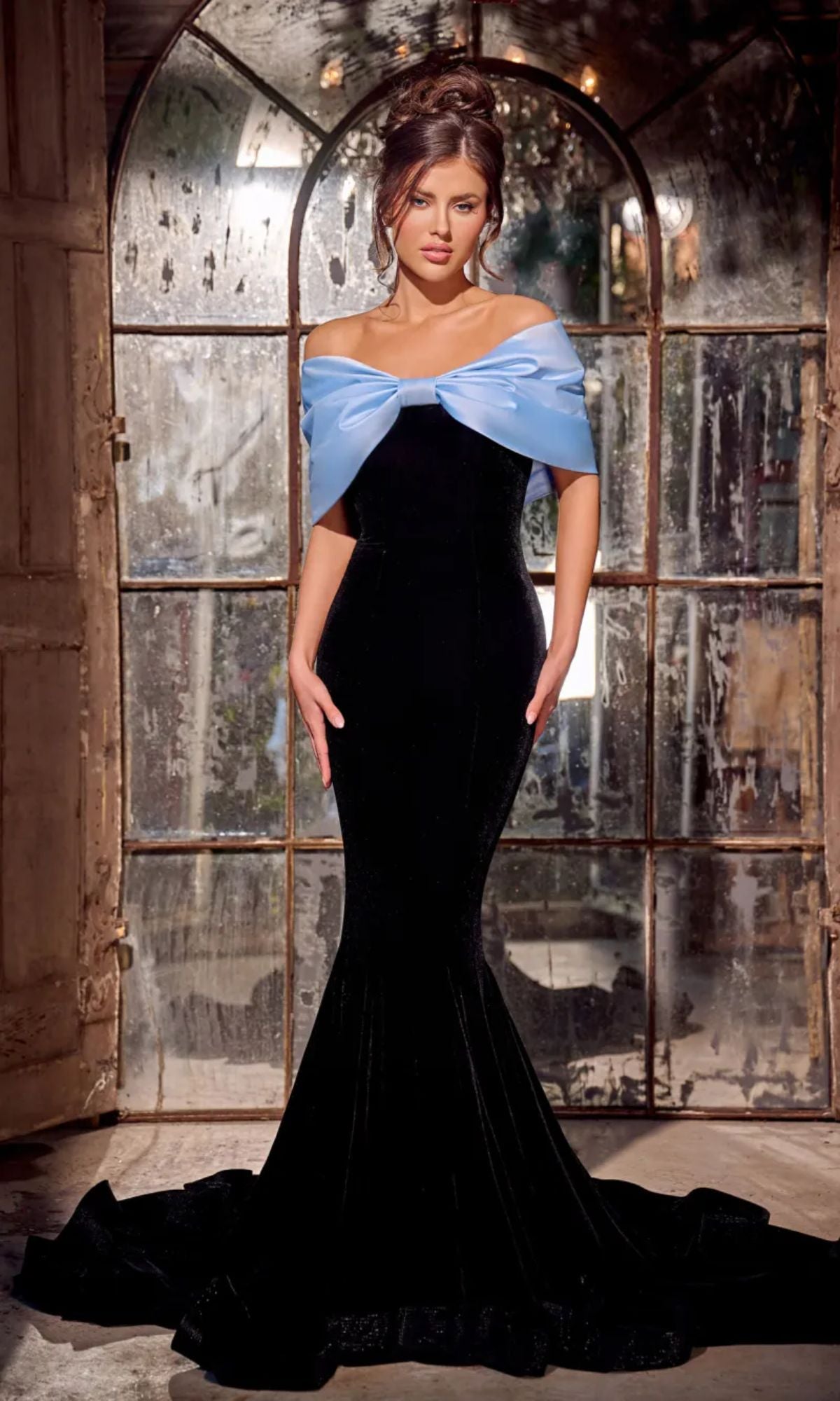 A woman stands indoors by an arched window wearing the Long Prom Dress PS24042 by Portia and Scarlett, an off-the-shoulder mermaid gown featuring a large light blue bow.