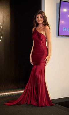 A woman wearing the Portia and Scarlett PS24051X long prom dress poses indoors by a lit display screen, smiling at the camera.