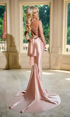 A woman with long blonde hair stands indoors, wearing the Long Prom Dress PS24091X by Portia and Scarlett— a fitted, shiny blush pink gown with a bow and open back. Through large windows behind her, lush greenery can be seen.