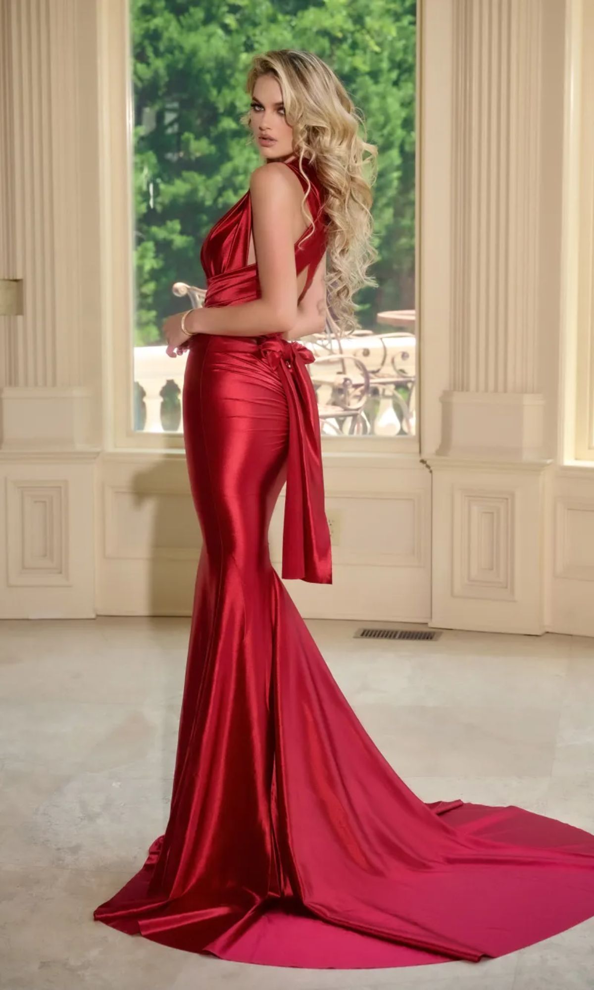 A woman with long, wavy blonde hair wears the Long Prom Dress PS24091X by Portia and Scarlett, featuring a shiny, fitted design with a plunging neckline and open back, standing indoors near large windows overlooking greenery.