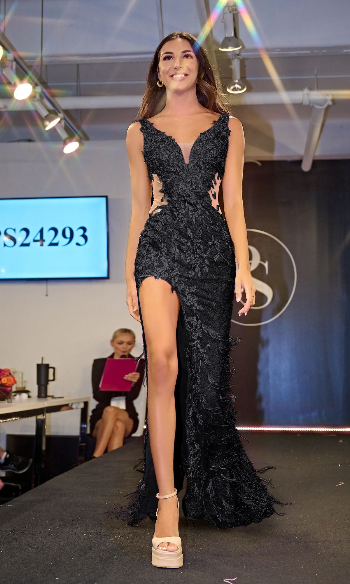 A woman walks the runway in the Long Prom Dress PS24293 by Portia and Scarlett, featuring intricate lace embroidery and a high slit; a judge with a pink clipboard sits in the background.