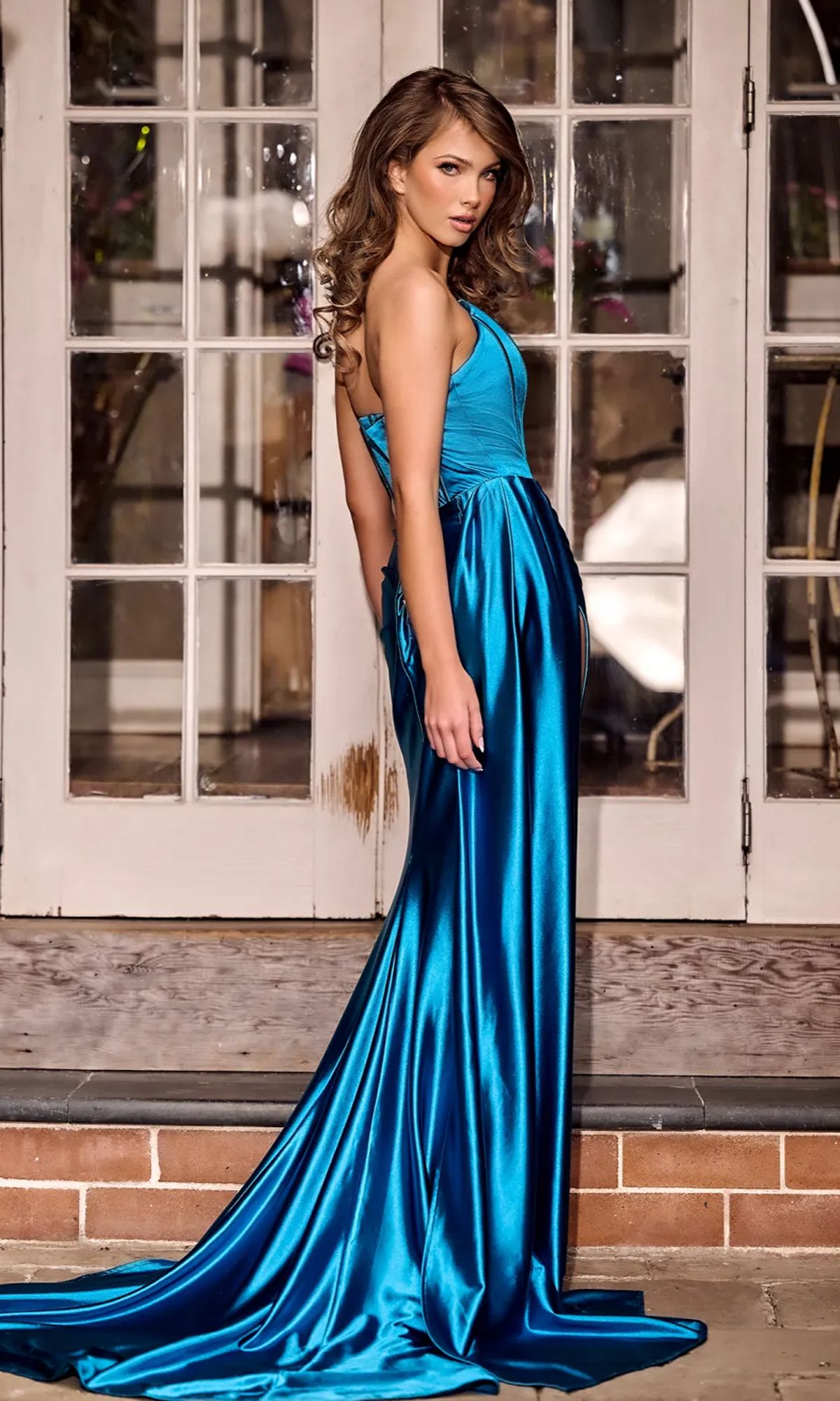 A woman in a shiny blue Strapless Satin Prom Gown: Portia & Scarlett PS24682 stands sideways on a wooden porch in front of glass-paneled doors.