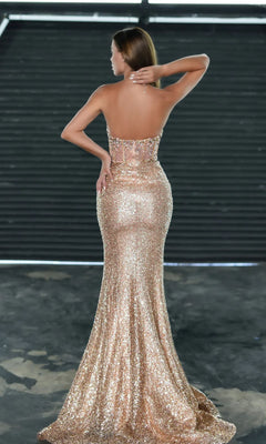 A woman faces away in an industrial setting, wearing the Portia and Scarlett Long Prom Dress PS24942—a strapless, floor-length, fitted corset dress with elegant gold sequins.
