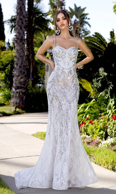 A woman stands outdoors among greenery and palm trees in the Portia and Scarlett PS25150 long prom dress, featuring a fitted ivory/silver lace design with thin straps and silver crystal beading.