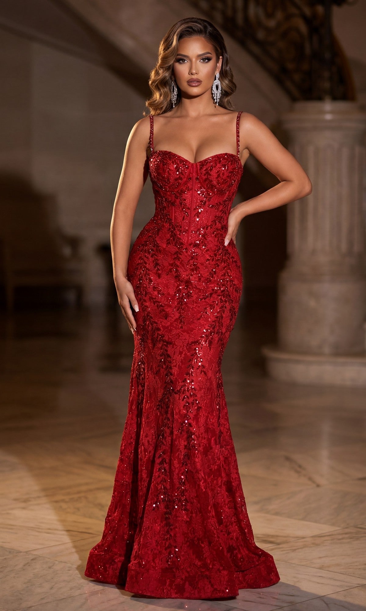 A woman stands indoors, posing with one hand on her hip in the fitted, floor-length red Portia and Scarlett PS25150 prom dress featuring sequins, a mermaid silhouette, thin straps, and a corset bodice.
