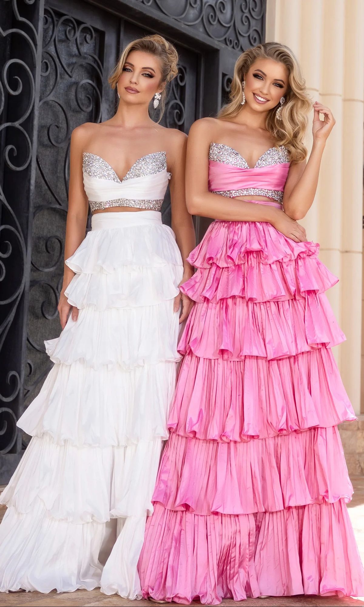Two women stand side by side in Portia and Scarlett PS25164 long prom dresses; both feature strapless, ruffled ball gown skirts with sparkling bodices—one dress is white, and the other is a bright pink.