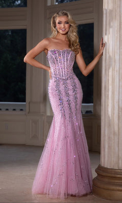 A woman wearing the Portia and Scarlett PS25169 long prom dress with a strapless lace mermaid silhouette and sparkling corset bodice stands indoors by a column, smiling at the camera.