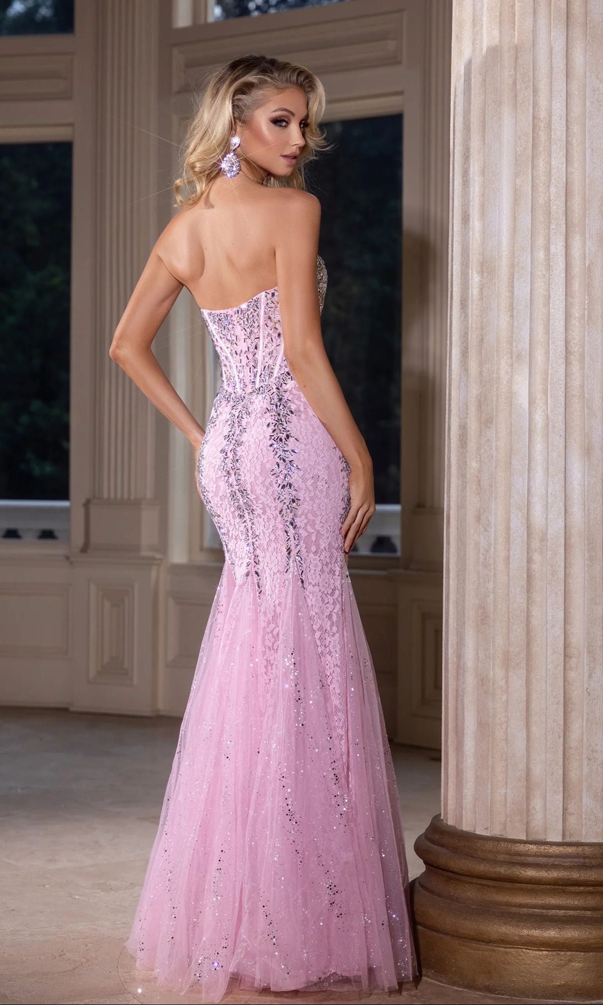 A woman stands indoors by a column, wearing the Portia and Scarlett PS25169 long prom dress—strapless, fitted, floor-length pink with sequin details and a corset bodice—looking over her shoulder.