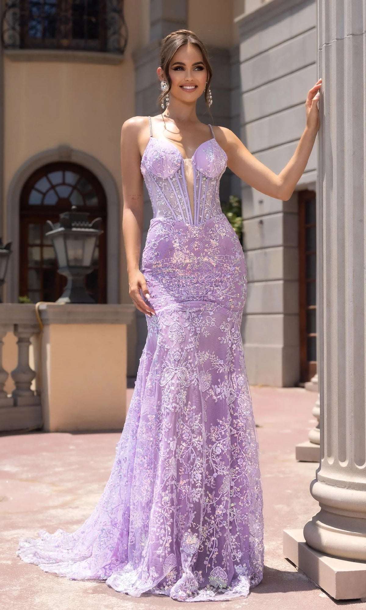 Wearing the Portia and Scarlett PS25234 long lilac prom dress with lace beaded embellishments and a sheer corset bodice, a woman stands outdoors smiling, her hand resting on a column.