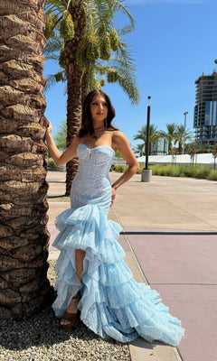 A woman wearing the Portia and Scarlett PS25250 long formal dress in blue, featuring a strapless boned corset bodice and ruffled tiered skirt, stands by a palm tree on a sunny day with modern buildings under a clear blue sky.