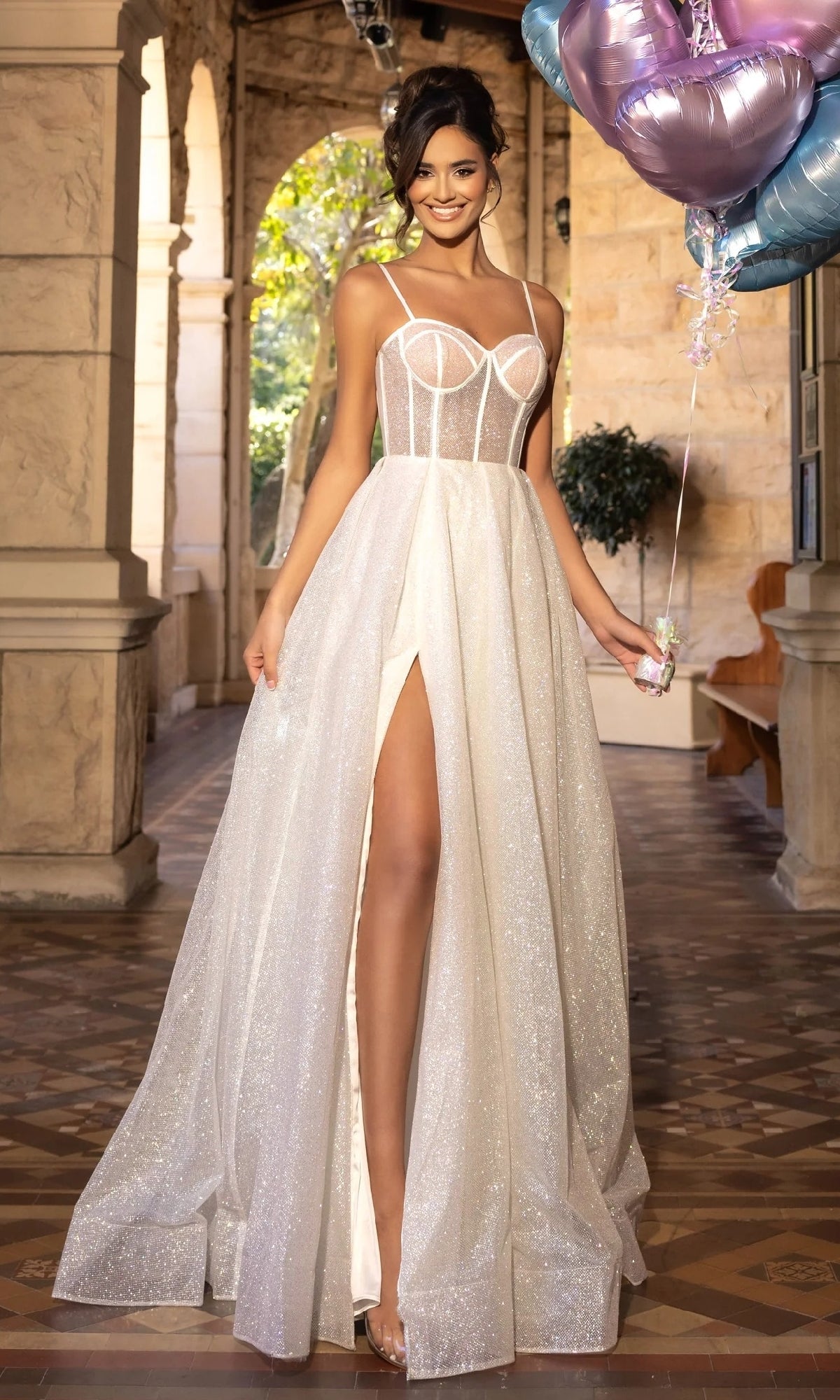 A woman in an ivory Portia and Scarlett PS25312 long prom dress with a sheer corset bodice and high slit stands indoors, holding balloons and smiling.